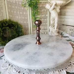 May include: A round marble cake stand with a copper-colored handle. The stand is white with gray veining and sits on a white crocheted doily. The handle has a decorative, textured top. The stand is ideal for displaying cakes or other desserts.