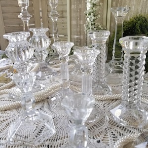 May include: A collection of clear glass candle holders of varying heights and designs. Some have faceted stems, while others have smooth or textured surfaces. The candle holders are arranged on a white lace tablecloth, with pearl strands.