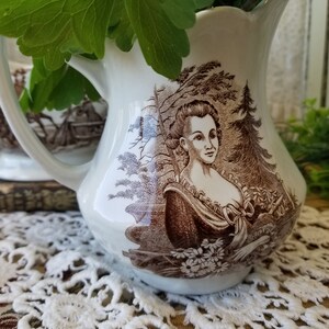 May include: A white ceramic creamer with a brown and white floral design. The creamer features a woman's portrait in the center.