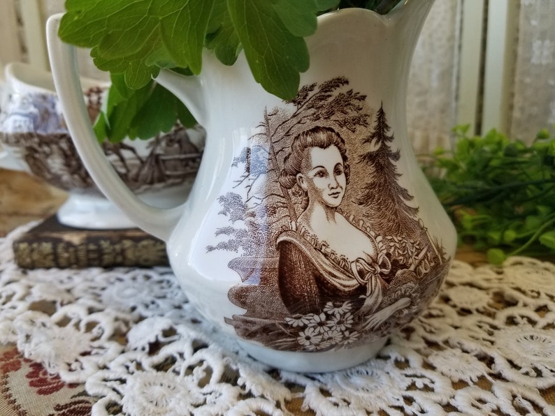 May include: A white ceramic creamer with a brown and white floral design. The creamer features a woman's portrait in the center.