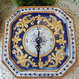 May include: A decorative ceramic clock with a blue, yellow, and white floral design. The clock face is surrounded by a blue border with a yellow and white floral pattern. The clock hands are black.