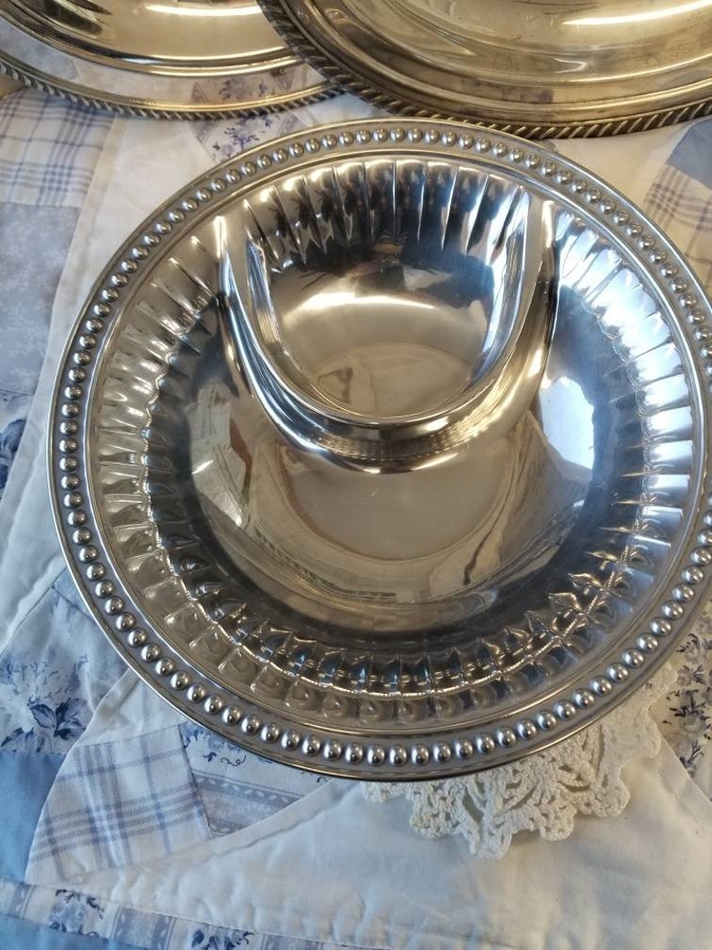 Puede incluir: Un conjunto de fuentes plateadas. Cada fuente tiene un compartimento central elevado y un borde con cuentas. Las fuentes son redondas y reflejan la luz, lo que sugiere una superficie pulida. El fondo es una tela estampada en azul claro y blanco.