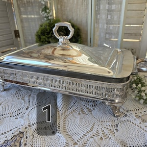 May include: A rectangular silver-plated chafing dish with a removable lid, featuring an ornate handle on top, two side handles, and four decorative feet. The base is embellished with a detailed embossed border pattern.