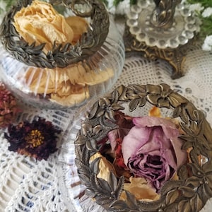 May include: Two clear glass bowls with vertical ribbing, each topped with an ornate, circular antique brass lid featuring a detailed leaf and floral relief pattern. The bowls are filled with dried flower petals, including yellow and purple roses.