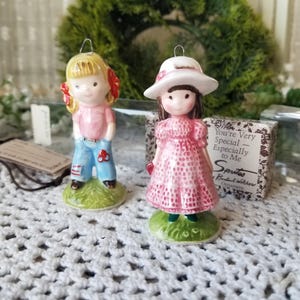 May include: Two ceramic figurines standing on green bases. One figure has blonde hair with red bows, a pink shirt, and blue jeans. The other figure wears a white hat and a pink patterned dress. A box nearby reads: You're Very Special — Especially to Me Sprites.
