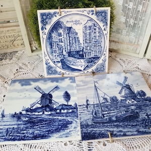 May include: Three blue and white ceramic tiles with Dutch scenes. One tile features a canal scene with the text "Amsterdam Holland". The other two tiles depict windmills and sailboats. The tiles are displayed on gold stands.