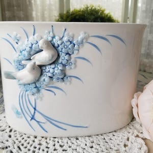 May include: White ceramic planter with a cylindrical shape, adorned with blue floral and bird relief details. The planter features a delicate design of blue flowers and two white birds, creating a charming, decorative piece.