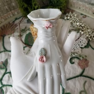 May include: A white porcelain vase in the shape of a hand, with a small bowl at the wrist. The vase is decorated with pink roses and gold trim. A decorative piece for the home.
