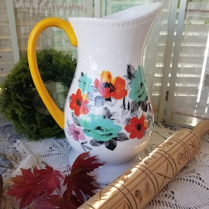 May include: White ceramic pitcher with a yellow handle, adorned with colorful floral patterns in red, orange, and teal. A wooden rolling pin with carved designs rests nearby, suggesting a kitchen or dining theme.