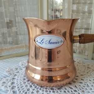 May include: A copper coffee pot with a wooden handle and a spout. The pot has a label that reads "Le Saucier" in blue script. The pot is sitting on a white crocheted doily.