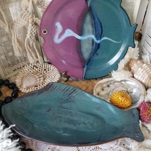 May include: Two ceramic fish-shaped serving dishes. One is a large plate with a pink and teal color scheme, and a white wave design. The other is a teal-colored platter. Decorative seashells and flowers are in the background.