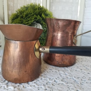 May include: Two copper coffee pots with black handles. The smaller pot has a gold accent on the handle. The pots are sitting on a white lace tablecloth.