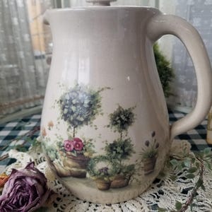 May include: A beige ceramic pitcher with a lid and handle, decorated with a garden scene featuring potted plants and flowers. The pitcher is set on a lace doily, with a dried rose and greenery nearby.