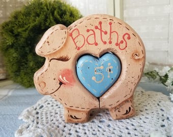 May include: A whimsical, wooden piggy bank in the shape of a pig. The bank is painted in a light peach color with the word "Baths" in red script. A blue heart-shaped coin slot is in the center, with the number "5¢" painted on it.