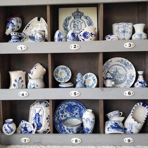 May include: A wooden display shelf filled with various blue and white ceramic pieces. The collection includes vases, plates, cups, and decorative figurines, all featuring intricate patterns and designs. The shelf is painted gray and has nine compartments.