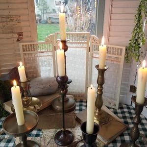 May include: Several lit ivory candles in various antique brass and bronze candle holders. The candles are of different heights and shapes, creating a warm, inviting ambiance. The background includes a white folding screen and a checkered tablecloth.