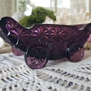 May include: A deep purple pressed glass decorative piece shaped like a vintage carriage or sleigh. The glass features a prominent starburst or daisy pattern across its surface and includes four circular wheels attached to the base of the body.