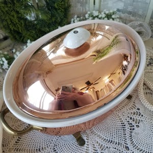 May include: Oval copper-colored serving dish with a white ceramic rim and lid. The lid has a white knob. The dish has brass handles and feet. The copper surface reflects the surroundings. A decorative piece for serving food.