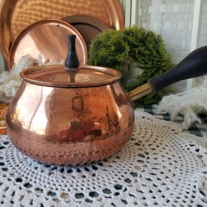 May include: A copper saucepan with a black wooden handle and lid. The pot is round with a shiny finish, reflecting the surrounding objects. The handle is attached with brass hardware. Other copper items are in the background.