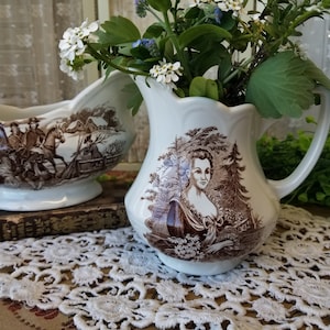 May include: Two vintage ceramic creamer pitchers with brown and white floral designs. The pitcher on the left features a scene of people riding horses. The pitcher on the right features a woman in a brown dress.