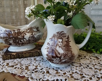 May include: Two vintage ceramic creamer pitchers with brown and white floral designs. The pitcher on the left features a scene of people riding horses. The pitcher on the right features a woman in a brown dress.