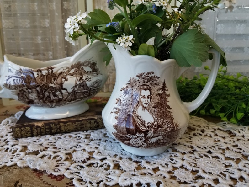May include: Two vintage ceramic creamer pitchers with brown and white floral designs. The pitcher on the left features a scene of people riding horses. The pitcher on the right features a woman in a brown dress.