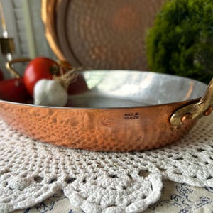 May include: An oval hammered copper pan with two brass handles riveted to the sides. The interior is lined with silver-colored metal. The exterior features a stamped mark that reads MADE IN FRANCE. The pan has a textured, dimpled surface finish.