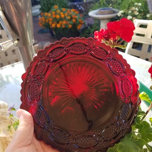 May include: A ruby red glass plate with a scalloped edge and intricate embossed detailing. The plate has a sunburst pattern in the center and is held in a hand. The plate is approximately 8 inches in diameter.