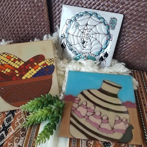 May include: Three decorative ceramic tiles with Southwestern designs. One tile features colorful corn in a brown bowl, another a dreamcatcher, and the third a vase with figures. The tiles are on a patterned surface.