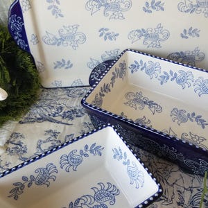 May include: A set of white ceramic baking dishes with a blue floral pattern. The dishes have a rectangular shape with rounded corners and blue rims. The pattern includes stylized flowers and leaves. The set includes a large rectangular dish and two smaller ones.
