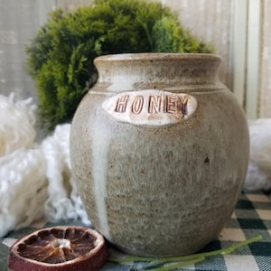 May include: A rustic, ceramic honey pot with a textured, speckled finish. The word "HONEY" is embossed on an oval label. The pot is a muted green-gray color, with a vertical white streak. A dried orange slice and greenery are in the foreground.