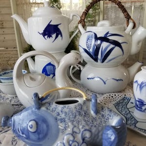May include: A collection of white ceramic teapots and serving dishes with blue painted designs. The teapots feature various shapes, including a fish-shaped teapot, and are decorated with fish, wheat, and wave motifs.