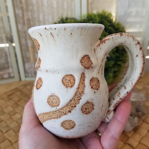 May include: Cream-colored ceramic mug with brown polka dots and a curved line design. The mug has a rounded body, a sturdy handle, and a slightly flared rim. The handmade mug is a unique piece of pottery.