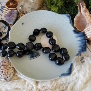 May include: A black beaded necklace rests on a light blue fish-shaped plate with dark blue accents. The plate sits on a white, textured surface, with seashells and greenery in the background. A dried orange slice and crystals are also visible.