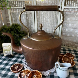 May include: A large, round, hammered copper tea kettle with a brass handle and a matching lid. The kettle features a curved spout and a wooden grip on the handle. A small white ceramic creamer with a green floral pattern sits next to the kettle.