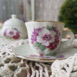 May include: A white teacup and saucer set with a floral design in shades of purple, pink, and green. The cup and saucer have gold trim. A matching teapot is slightly out of focus in the background. The set is on a white lace tablecloth.