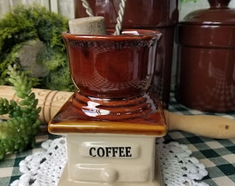 May include: A ceramic coffee grinder replica featuring a glossy brown top section and a cream-colored base with the word COFFEE printed in black lettering. A metal wire whisk is positioned inside the top opening of the grinder.