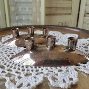 May include: Two silver-toned candle holders, each with three cylindrical cups, sit on a silver tray atop a white crocheted doily. The oval-shaped holders have a reflective surface, and the background is softly blurred.