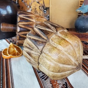May include: A carved wooden decorative object with a textured surface, resembling a fruit or nut. The object is light brown with darker brown accents. Two dried orange slices are visible. The background includes a jug and a decorative wooden rolling pin.