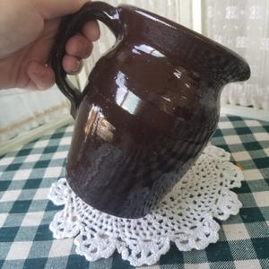 May include: A dark brown ceramic pitcher with a handle. The pitcher is glossy and appears to be handmade. It sits on a white, crocheted doily over a green and white checkered tablecloth.