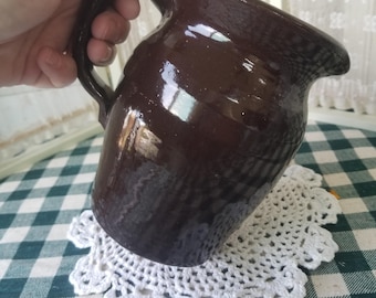 May include: A dark brown ceramic pitcher with a handle. The pitcher is glossy and appears to be handmade. It sits on a white, crocheted doily over a green and white checkered tablecloth.