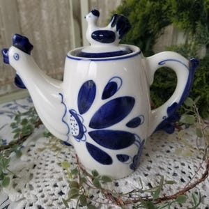 May include: White ceramic teapot with blue floral and rooster design. The teapot features a rooster-shaped spout and a rooster figurine on the lid. The handle and accents are also blue. The teapot is sitting on a white lace doily.