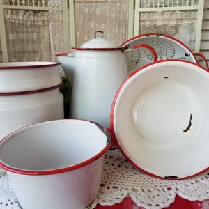 May include: A collection of white enamelware kitchen items with red trim. The set includes a large pot, a colander, a bowl, and a small container. The items are arranged on a red and white checkered tablecloth, evoking a vintage aesthetic.