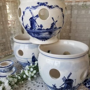 May include: Three white ceramic bulb planters with blue windmill and landscape designs. Each planter has a series of circular holes. The planters are stacked on top of each other. A small ceramic dish and a sprig of white flowers are also visible.