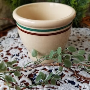 May include: A small, cream-colored ceramic cup with a brown and green striped band around the rim. The cup is sitting on a white lace doily.