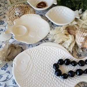 May include: White ceramic fish-shaped serving dishes and plates. The plates have a scale pattern and are arranged with a black beaded necklace. A seashell and quartz crystals are also present.