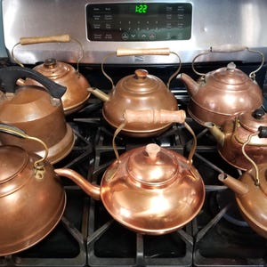 May include: A collection of copper teapots and kettles with wooden and black handles. The teapots vary in shape and size, with some featuring spouts and lids. The copper surfaces reflect light, creating a warm, inviting aesthetic.