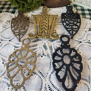 May include: A collection of five metal trivets in various shapes and finishes, including two black cast iron pieces, two brass-toned metal pieces with intricate openwork designs, and one rectangular brass-toned trivet with a central leaf motif.
