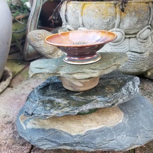 May include: A small, brown ceramic bowl with a flared rim sits atop a stack of gray and tan stones. The bowl has a glossy finish and a white base. The stones are arranged on a red brick surface, with a blurred background of plants and a decorative pot.