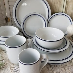 May include: A white ceramic dinnerware set featuring a simple dark blue band accent along the rims. The collection includes multiple round dinner plates, smaller side plates, deep bowls, and two mugs, all displaying a consistent minimalist design.